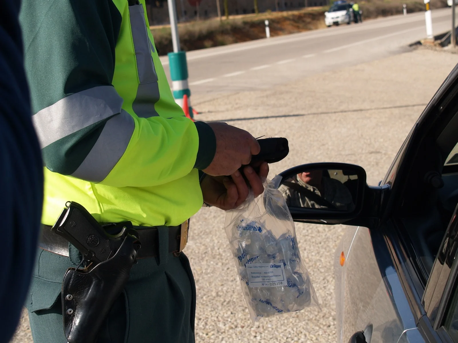 Police officer at a roadside stop holding a breathalyzer device and a bag of sterile mouthpieces beside a stopped car